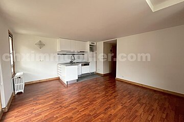 Empty studio apartment with white walls, reddish-brown wooden floor, and a modern white kitchenette. A window with a radiator is on the left.