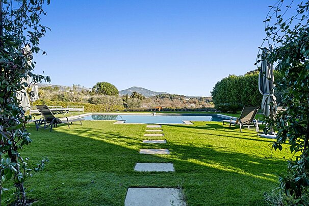 A lush backyard with a swimming pool, lounge chairs, a green lawn, stepping stones, and distant mountain views under a clear blue sky.
