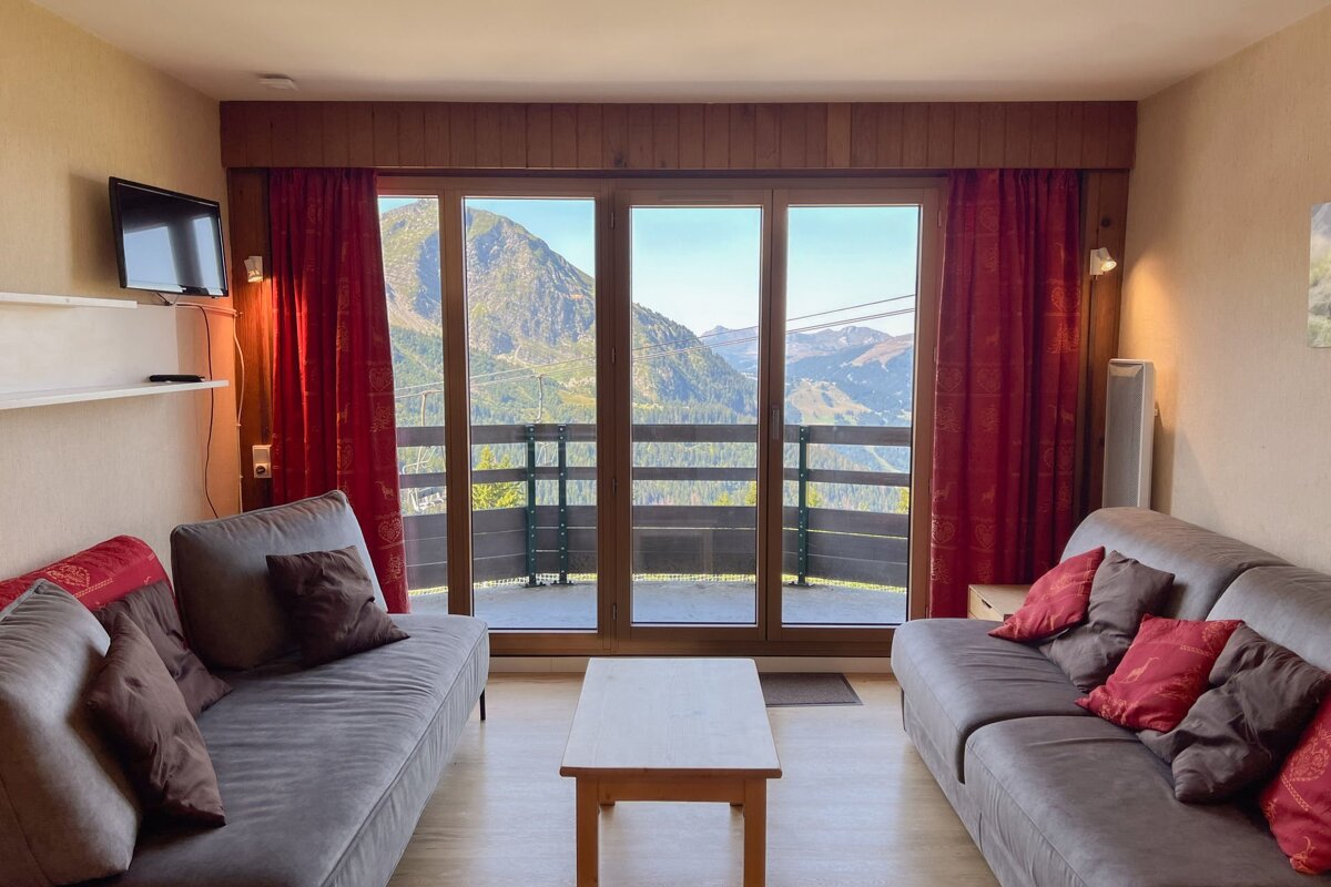 An inviting living room with two sofas, a coffee table, and large glass doors showcasing a panoramic mountain view. Red curtains accent the scenic outlook.