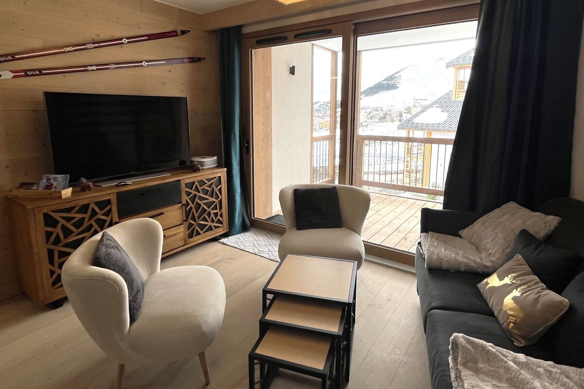 A cozy, modern living room with a TV, ski decor, and plush seating. Large glass doors open to a balcony with a snowy mountain and resort view.