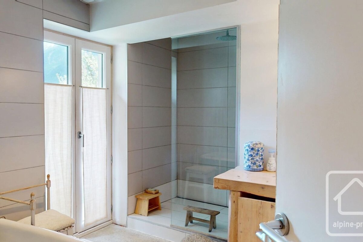 Bright bathroom with a walk-in shower (grey tiles, glass), double doors with blinds, wooden stools, and a vanity. A partial tub is on the left.