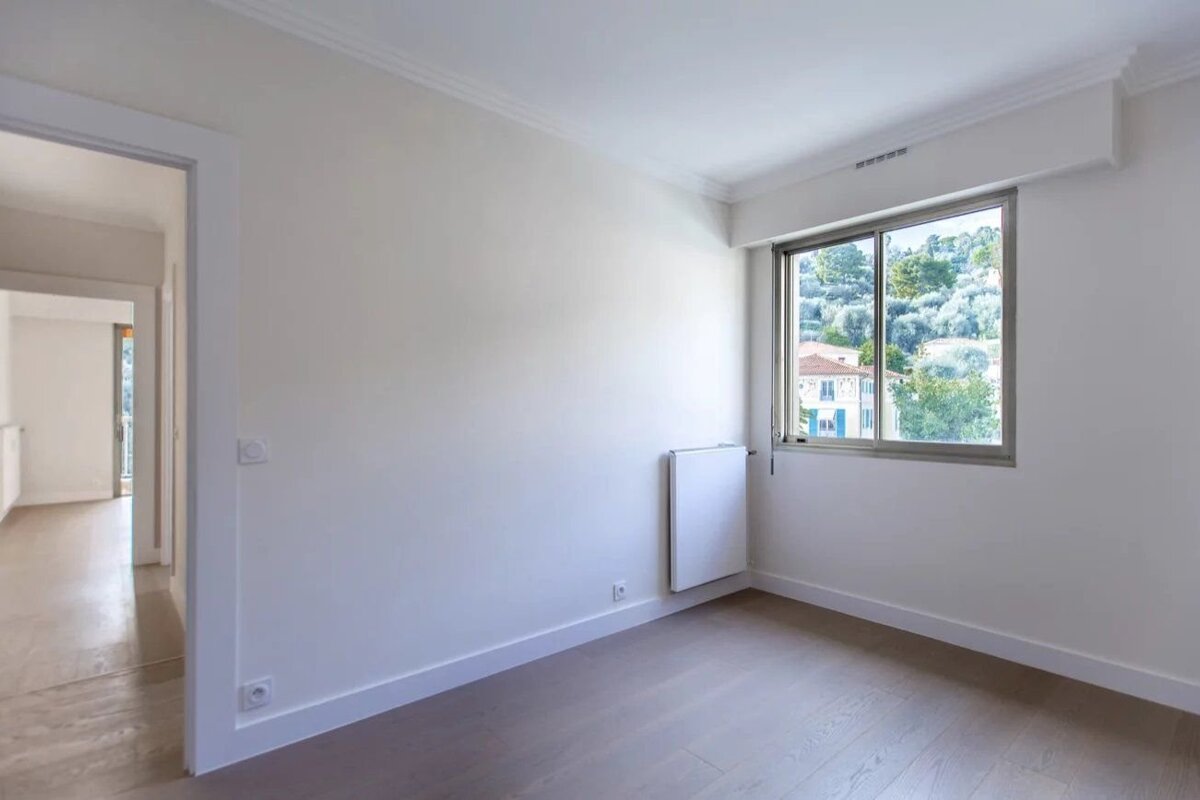 An empty modern room with white walls, light wood floors, an open doorway on the left, and a window showing a green outdoor view.
