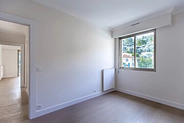 An empty modern room with white walls, light wood floors, an open doorway on the left, and a window showing a green outdoor view.