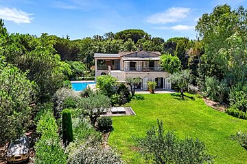 A grand villa with a pool and extensive green lawn, surrounded by lush trees under a blue sky. An old car is nestled in the garden foliage.