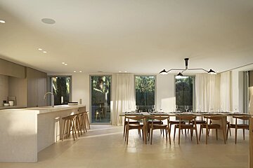 Modern kitchen and dining room with a long island, wooden dining table, and large windows revealing a lush outdoor view.