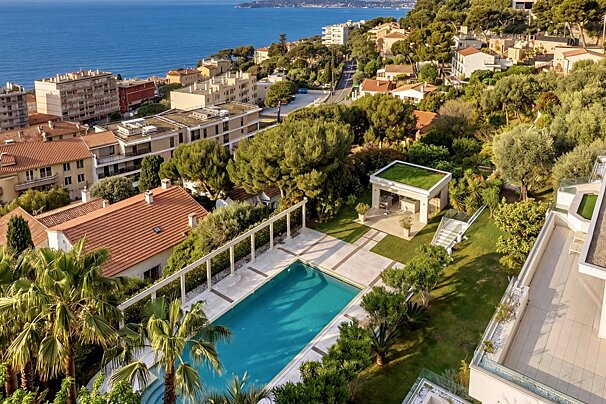 Drone view of a sunny coastal town featuring houses, a large swimming pool, green gardens, and the deep blue sea in the distance.
