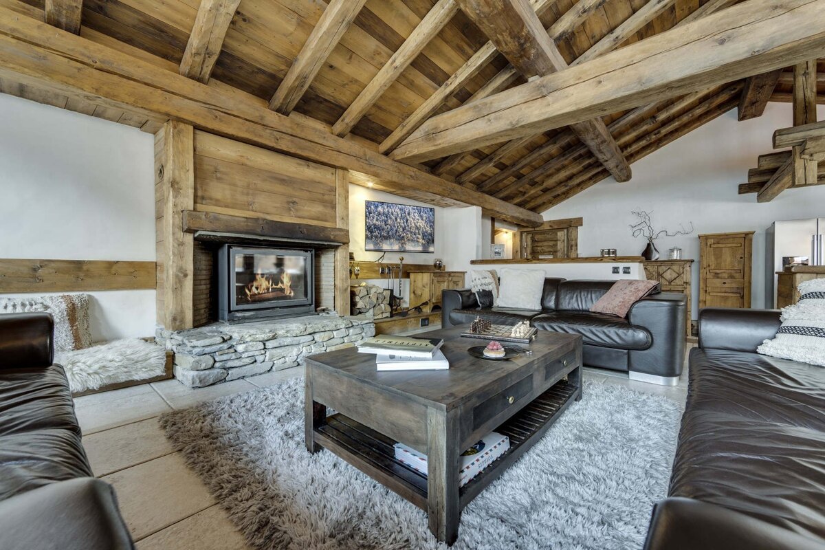 Cozy, rustic chalet living room featuring exposed wood beams, a stone fireplace with a fire, dark leather sofas, and a large coffee table.