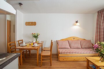 A bright, rustic open-plan living/dining room with a wooden table set for a meal, a plaid daybed, fresh flowers, and kitchen counter visible.