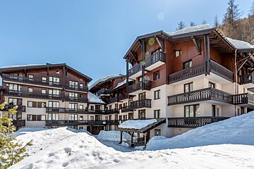 Chalet-style apartment buildings surrounded by deep snow under a clear blue sky, typical of a bright winter day at a ski resort.