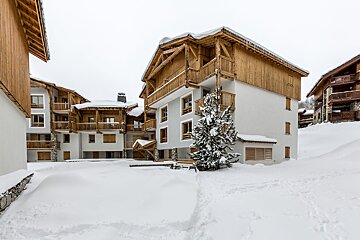 Snow-covered ski resort buildings with white walls and rustic wooden accents, featuring snow-laden roofs and a tree under an overcast winter sky.