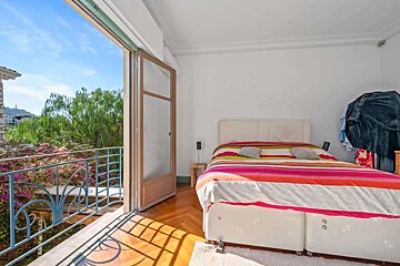 A bright bedroom with a striped bed, open doors to a balcony overlooking lush greenery, flowering plants, and a blue sky. Sunlight casts shadows on the wooden floor.