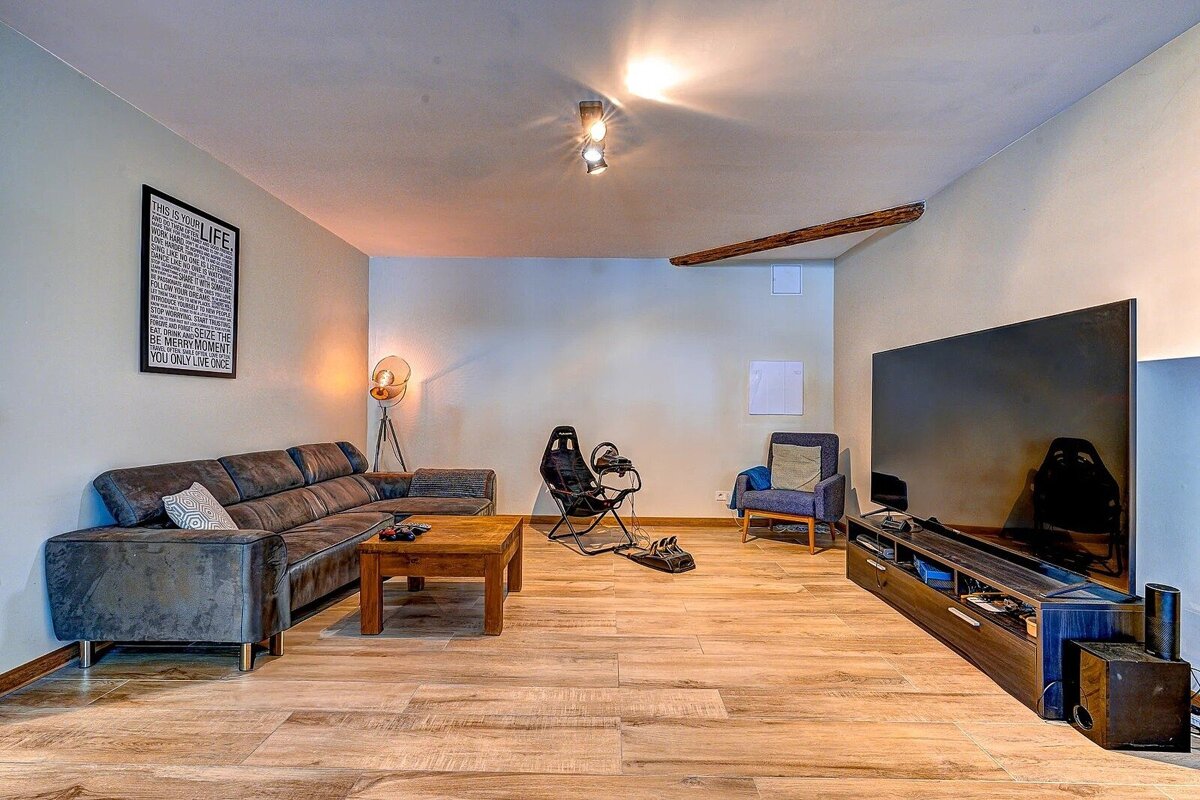 A spacious living room with a large sofa, wooden coffee table, big TV, and a racing sim setup. Warm lighting illuminates the wooden floor.