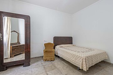 A simple bedroom with a single bed, a yellow armchair, and a large wooden mirrored wardrobe reflecting a dresser.