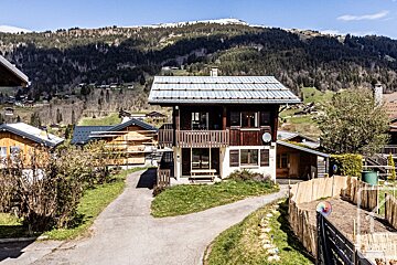 A cozy chalet-style house with a balcony stands in a green mountain village under a clear sky, surrounded by other homes and forested hills.