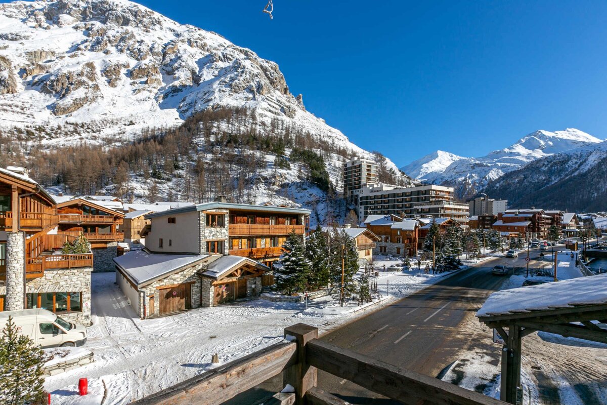 A snowy alpine village with wooden chalets and a road, set against majestic, snow-capped mountains under a clear blue sky.