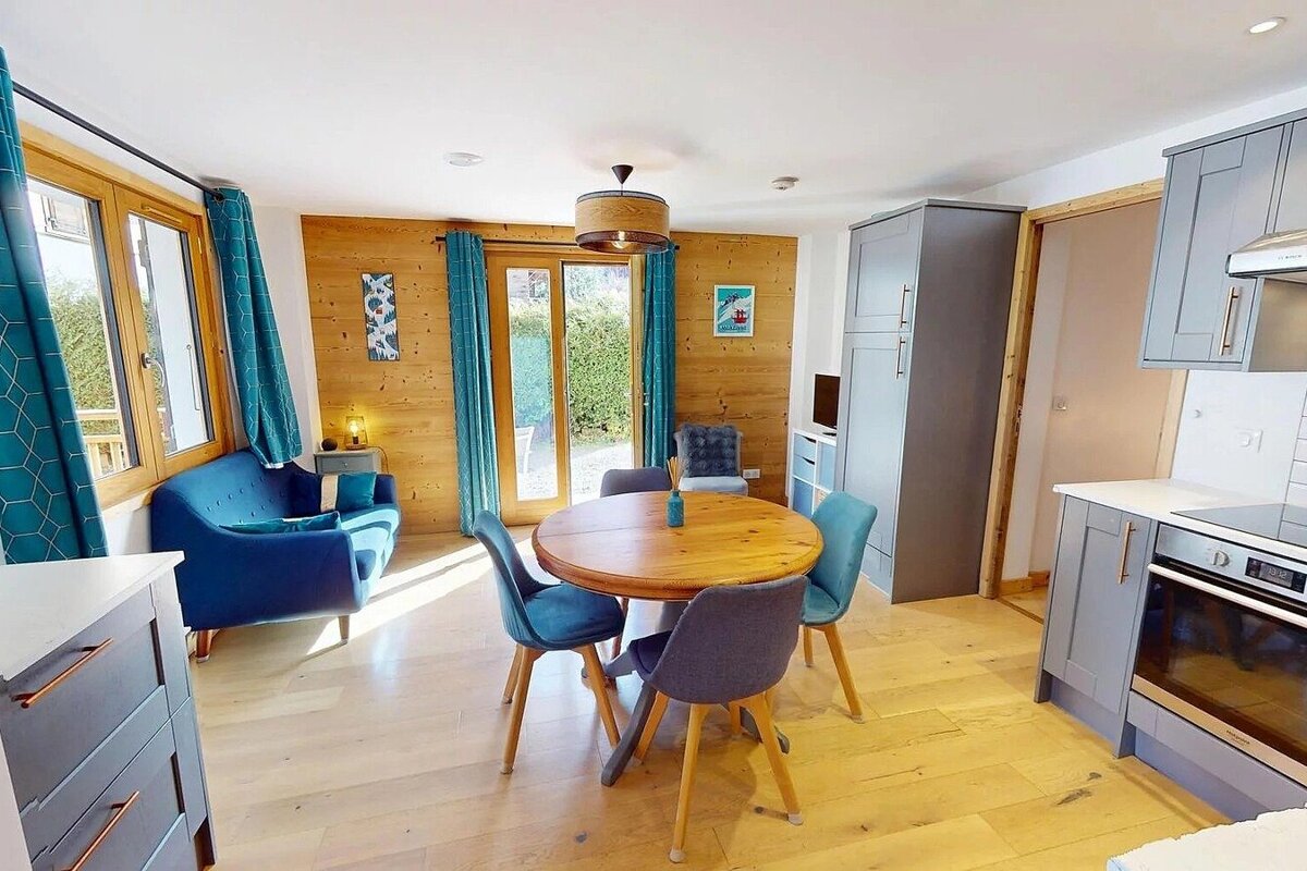 An inviting open-plan room with light wood floors & walls, a blue sofa, round dining table, and a modern grey kitchen. Teal curtains and chairs add color.