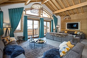 Cozy chalet living room with high wooden ceilings, grey sofas, colorful pillows, and teal curtains framing large windows.