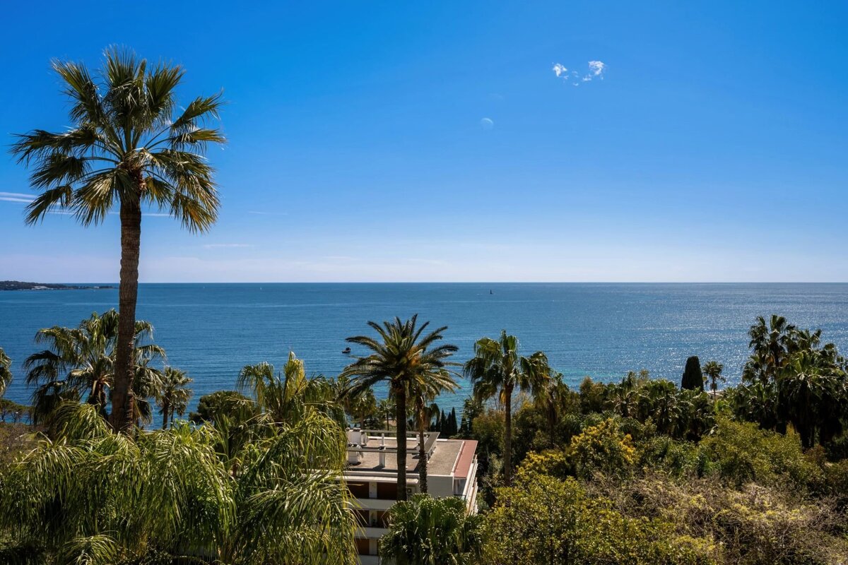 Palm trees frame a sparkling blue sea under a clear sky. Green foliage and a building are visible below.