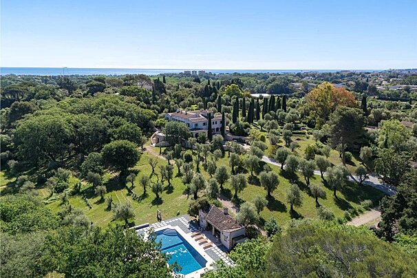 Aerial view of a luxury estate with a villa, lush green gardens, a swimming pool, and the sea on the horizon.