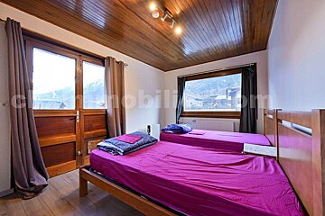 A rustic bedroom with a wood-paneled ceiling, two single beds with bright pink sheets, wooden floors, and large windows revealing a mountain village view.
