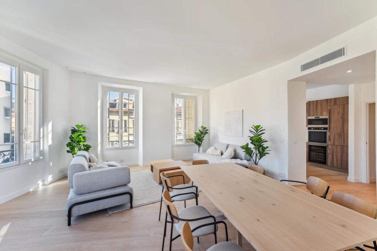Bright, modern living and dining area with light wood furniture, white walls, large windows, and an adjacent kitchen.