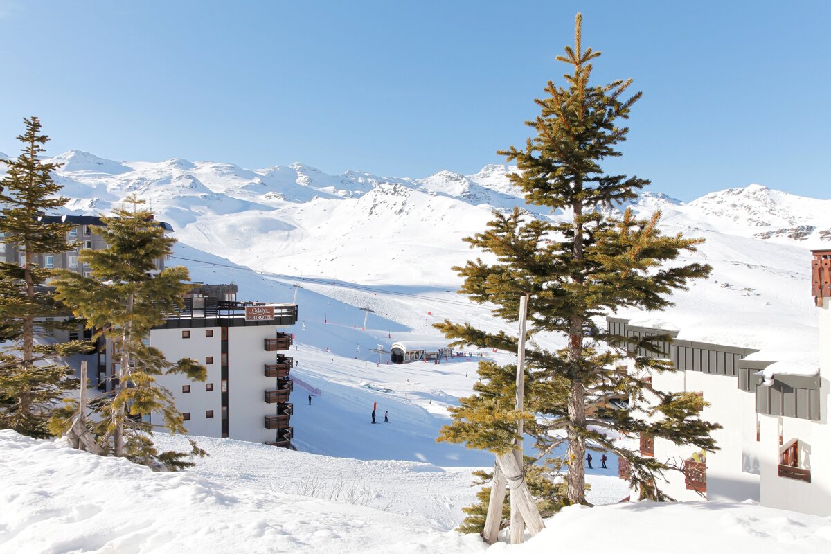 A bright, snowy mountain ski resort with buildings, pine trees, and skiers on the slopes, all under a clear blue sky.