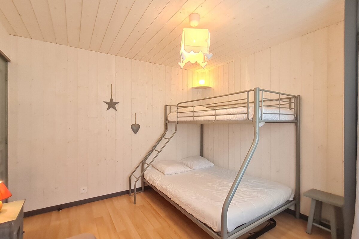 A light wood-paneled room features a metal bunk bed with white bedding, wooden floors, and hanging star/heart decor, illuminated by a warm ceiling light.