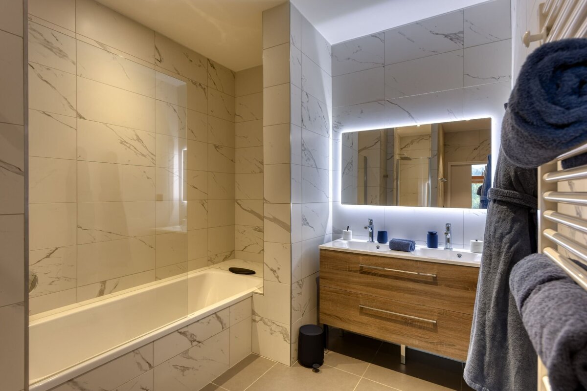Modern bathroom with white marble-look tiles, a bathtub, a wooden double vanity with an LED-backlit mirror, and blue towels nearby.