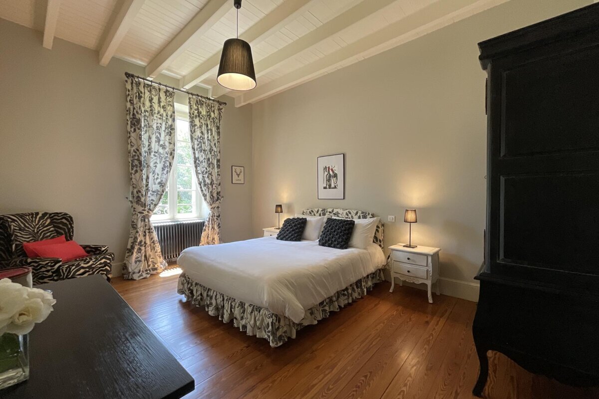 A spacious, elegant bedroom featuring a large bed with a patterned skirt, hardwood floors, high beamed ceilings, and a large window with patterned curtains.
