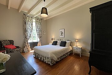 A spacious, elegant bedroom featuring a large bed with a patterned skirt, hardwood floors, high beamed ceilings, and a large window with patterned curtains.