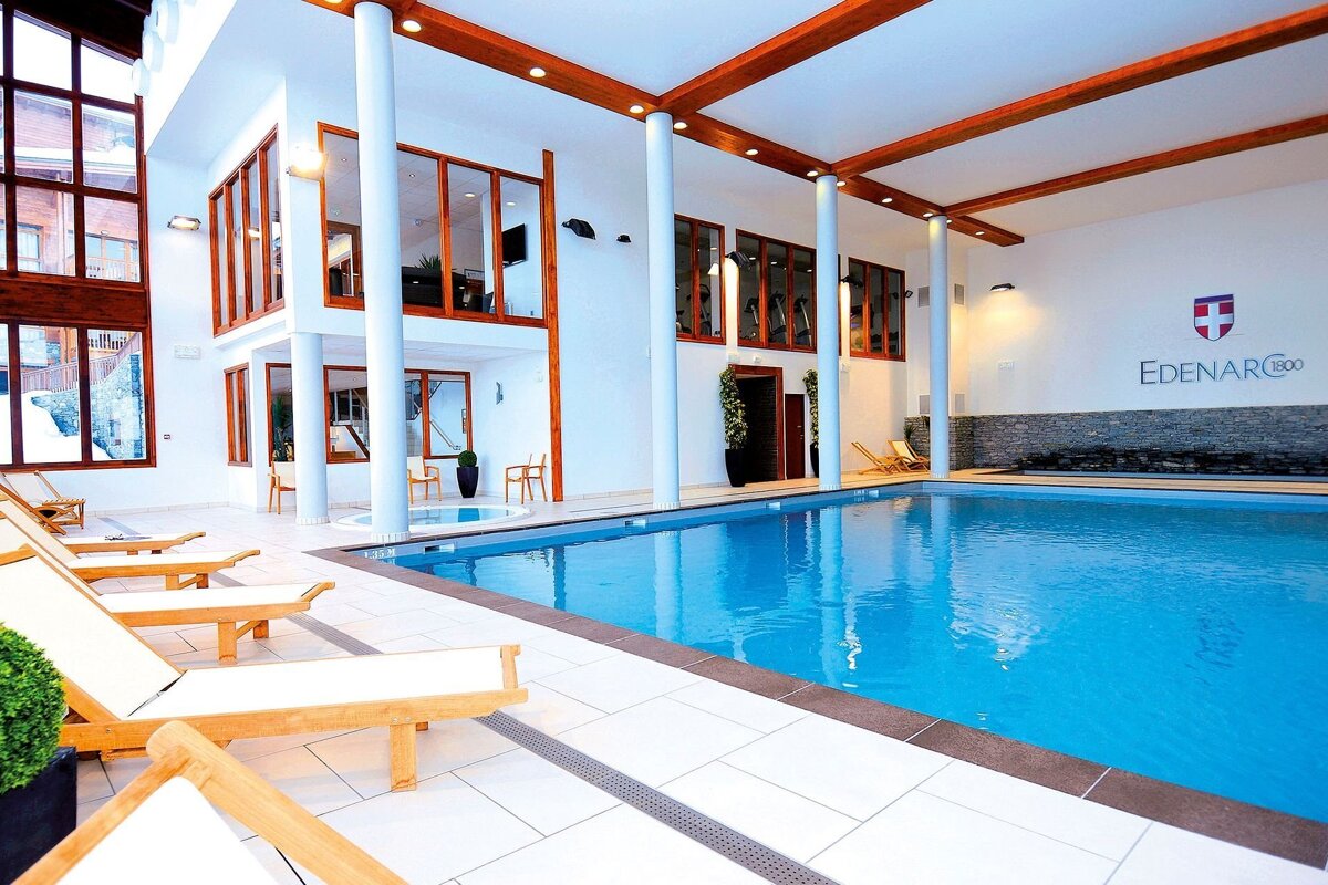 A bright, modern indoor swimming pool and hot tub area with lounge chairs, large windows, and a high ceiling with wooden beams. 