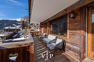 Sunny wooden balcony of an alpine chalet, featuring outdoor seating and stunning views of snowy mountains.