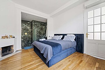 A modern bedroom with a blue bed, hardwood floor, white walls, an open shower with dark marble, a fireplace, and a glass-paned door.