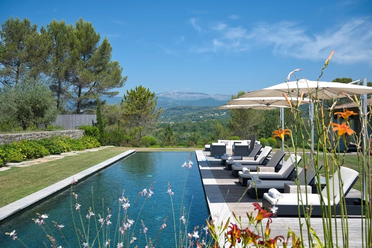 Luxury outdoor pool with sun loungers and umbrellas overlooking hills and mountains under a clear blue sky.