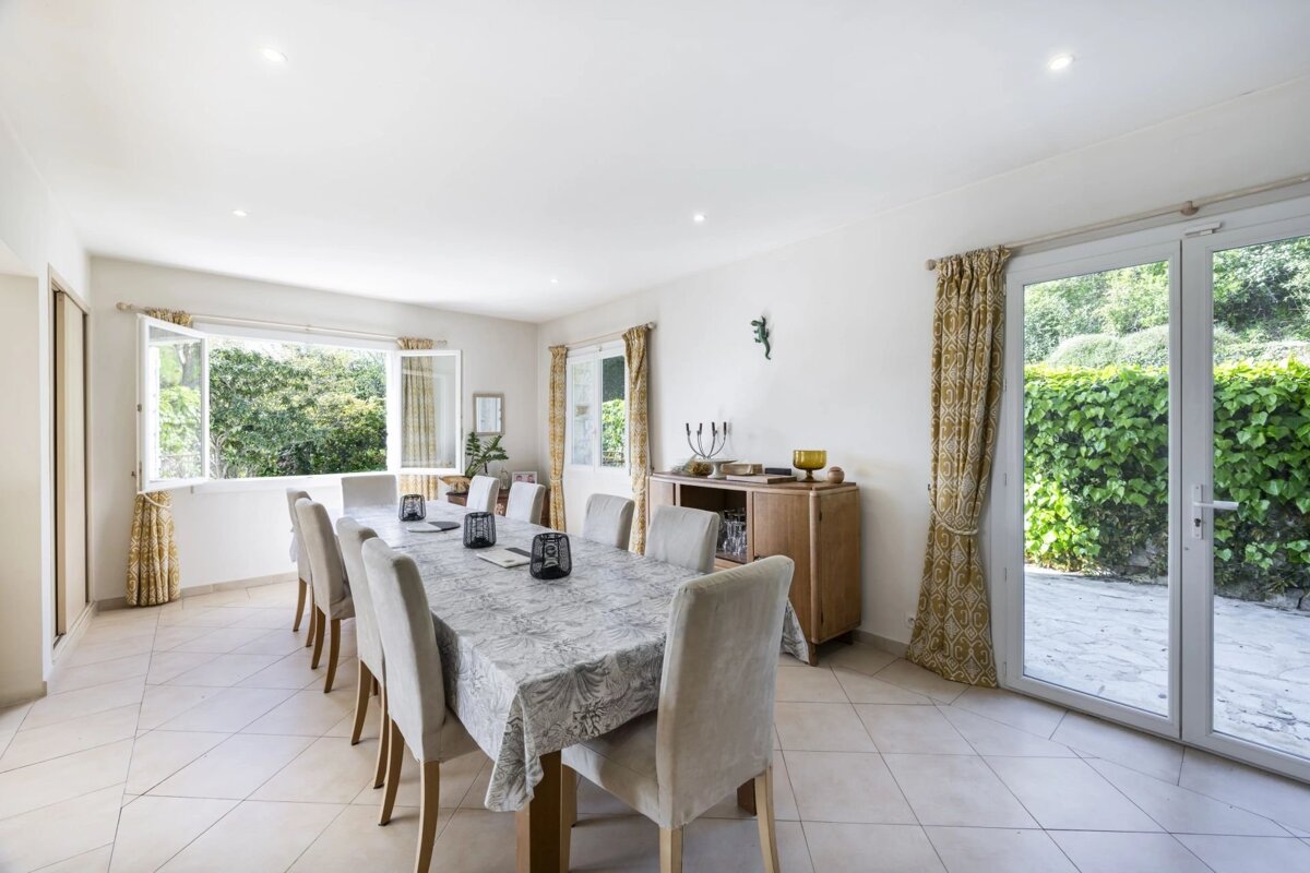 A bright, modern dining room features a long table with upholstered chairs, large windows revealing lush greenery, and a patio door to a garden.