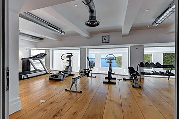 A bright, luxurious home gym with diverse exercise equipment, hardwood floors, and large windows offering a stunning water view.
