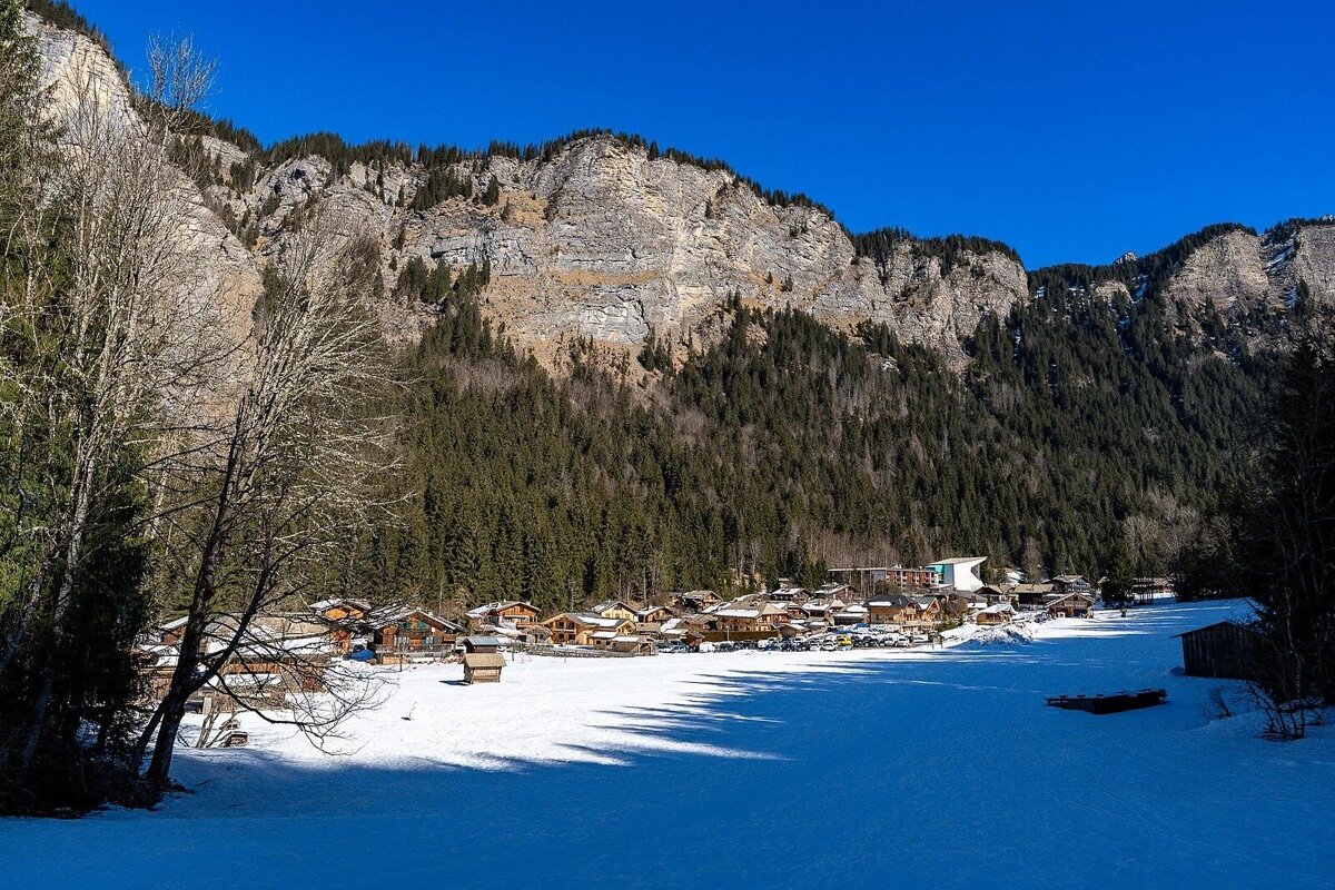 A snowy village nestled below a vast, forested mountain range under a clear blue sky.