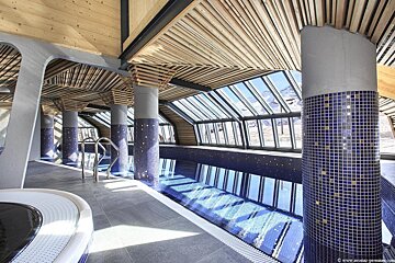 An indoor pool with blue mosaic-tiled pillars and a sloped glass roof showcasing views of snowy mountains.