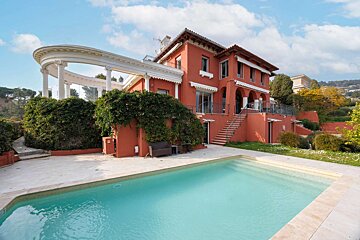 A vibrant red multi-story villa with a white columned balcony overlooking a bright blue swimming pool, set against a clear sky.