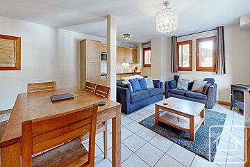 An open-concept living area with a wooden dining table, a tiled floor, two dark sofas, a coffee table, and a kitchen in the background.