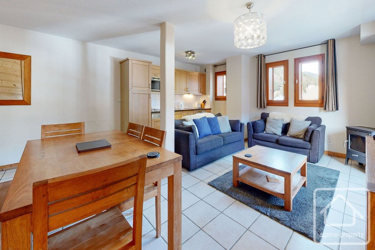 Bright, open-plan living and dining area with a wooden table, two blue sofas, a coffee table, and a glimpse of the kitchen. White tiled floor.