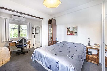 Bright bedroom with a floral bedspread, vintage desk by a window, wooden display cabinet, and a painting on the white wall.