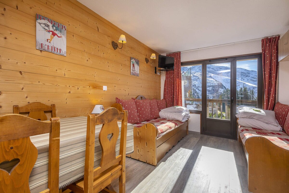 A cozy, wood-paneled chalet room with a dining table, two daybeds, and ski decor. A large window offers stunning views of snowy mountains.