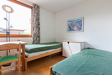A bright, simple bedroom with two single beds (green sheets), a desk, lamp, window with floral curtain, white cabinet, and a map on the wall.