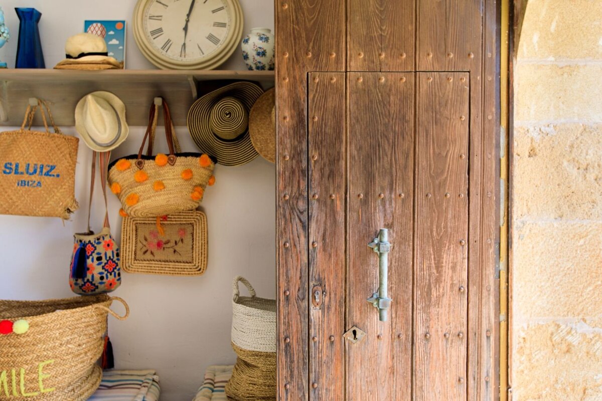 A rustic wooden door next to shelves displaying straw hats, bags, baskets, and a large clock, creating a warm, summery ambiance.