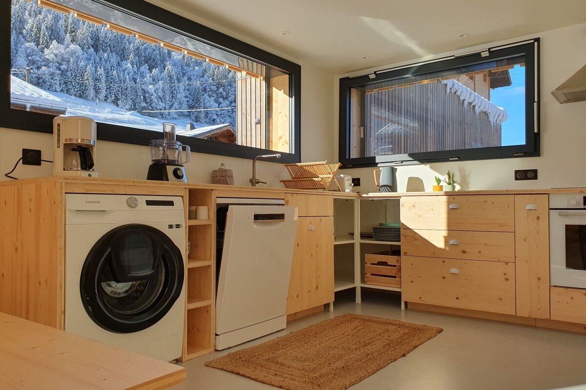 Sunny kitchen with light wood cabinets, washer, dishwasher, and large windows overlooking a snowy mountain landscape.