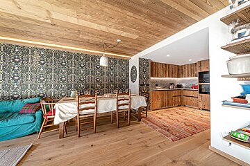 A cozy open-plan room with a dining area, turquoise sofa, and kitchen with wooden cabinets. Ornate wallpaper and wood textures create a rustic feel.