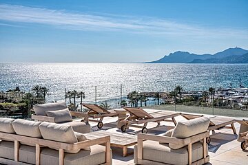 Modern patio with lounge furniture overlooking a sparkling blue sea, distant mountains, and a harbor with yachts under a clear sky.