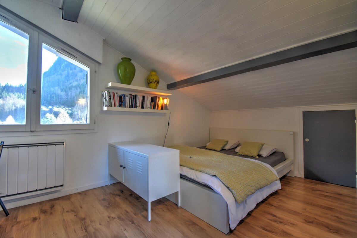 Cozy attic bedroom with wood floors, a bed with yellow bedding, a white cabinet, bookshelves, and a window with scenic views.
