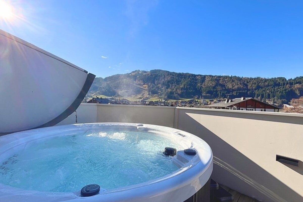 A steaming outdoor hot tub on a terrace with a beautiful view of forested mountains under a clear blue sky.
